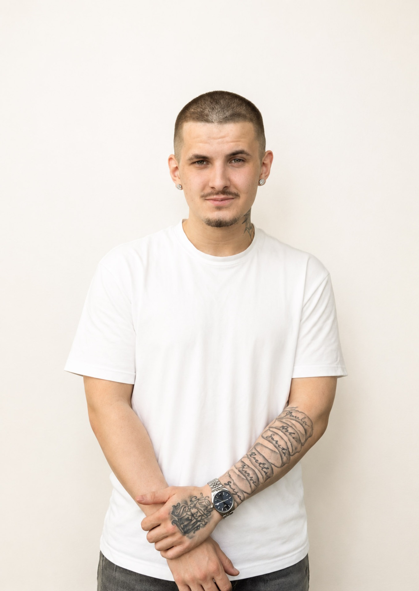Nuori mies valkoisessa t-paidassa, lyhyet hiukset, tatuoitu käsivarsi ja taustalla vaalea studio.